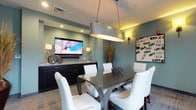 This single-floor space exudes a contemporary elegance through its thoughtfully curated design and refined finishes. The centerpiece of the room is a sleek, dark rectangular dining table surrounded by plush white upholstered chairs, creating an inviting setting for gatherings or meetings. Above the table, a striking oval metallic pendant light suspended by chains serves as a bold focal point, casting warm, ambient light throughout the space. The walls are painted in a soothing muted teal-green tone, contributing to the room's calm and sophisticated atmosphere. Flanking the walls, warm sconce lighting adds layers of illumination that enhance the overall ambiance. A built-in dark wood credenza lines one wall, topped with a granite or stone surface and anchored by a large flat-screen TV mounted above — making the space as functional as it is stylish. Decorative elements, including sculptural wire orbs and tall ornamental grasses in a vase, add texture and visual interest throughout. Framed artwork on the wall completes the polished aesthetic. Recessed ceiling lighting and a neutral-toned carpet floor tie the design together, resulting in a versatile, upscale space well-suited for dining, entertainment, or private events.
