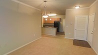 This single-floor residence offers an open-concept layout that integrates the living area and kitchen into one cohesive space. The living area is finished with plush, neutral-toned carpeting and crisp white baseboards, creating a clean and welcoming atmosphere. Crown molding lines the ceiling throughout, adding a touch of classic architectural detail.

The kitchen is positioned at the rear of the open layout and features a raised breakfast bar that serves as a natural divider between the cooking and living spaces. Warm honey-toned wood cabinetry provides ample storage, while dark countertops offer a contrasting, contemporary accent. A black refrigerator and built-in sink complete the functional kitchen setup. Overhead, a combination of recessed lighting and a decorative pendant light illuminate the bar area, while a stylish hanging pendant light fixture anchors the dining or transitional zone just outside the kitchen.

A hallway visible beyond the kitchen leads to additional areas of the unit, and a white paneled door on the right side of the living area provides access to a closet or utility space. The overall ambiance is understated and move-in ready, with neutral tones throughout.