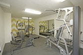 This single-floor fitness room offers a functional and well-equipped workout space designed to accommodate a variety of exercise routines. The room features dark rubberized flooring, which provides durability and traction suitable for physical activity. Overhead fluorescent lighting ensures the space is well-lit throughout.

The interior design incorporates a distinctive yellow and white chevron-patterned accent wall, adding a bold visual focal point to the room. Additional walls are painted in a clean white, while a solid yellow accent wall near the entry door contributes to the energetic color scheme. A textured ceiling completes the room's finish.

Equipment includes a multi-function cable weight station, a treadmill, an elliptical trainer, and a recumbent stationary bike, offering options for both strength training and cardiovascular exercise. A large wall-mounted mirror spans one side of the room, visually expanding the space and allowing users to monitor their form during workouts.

The room is accessed through a paneled white door and includes a floor vent for climate control. The overall layout is efficiently organized, maximizing usable floor space while keeping equipment accessible.