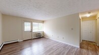 Located at 1908 Newton St in Akron, Ohio, this single-floor, one-bedroom, one-bathroom unit offers a practical and well-laid-out living space. The living room is generously proportioned, featuring light-toned luxury vinyl plank flooring, neutral beige walls, a textured ceiling, and baseboard heating. A wall-mounted air conditioning unit and a paneled entry door with an adjacent window allow for natural light and ventilation. A pass-through opening connects the living area to the kitchen, enhancing the sense of openness between the two spaces. The kitchen is equipped with warm oak cabinetry, a white electric coil-top range, and matching light flooring, providing a functional cooking environment. The bedroom offers a comfortable, carpeted floor, a textured ceiling, baseboard heating, a window for natural light, and a closet for storage. The full bathroom features a white soaking tub with an overhead shower, white square wall tiles, a toilet, and wood-look flooring, presenting a clean and utilitarian design. Throughout the unit, the neutral color palette and consistent finishes create a cohesive and easy-to-personalize living environment.