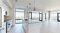 This contemporary single-floor residence offers a sleek, open-concept layout designed to maximize natural light and panoramic views. The living and kitchen area form the heart of the home, anchored by a large central island with a deep undermount sink, quartz countertops, and white flat-front cabinetry. Pendant lighting in muted concrete tones adds a modern touch overhead, while floor-to-ceiling windows with black steel frames frame expansive views of the city skyline and waterfront beyond. Light hardwood flooring flows seamlessly throughout, enhancing the bright, airy atmosphere. Multiple rooms open directly onto outdoor terraces via full-height glass doors, blurring the line between interior and exterior living. The bedrooms are generously proportioned, each benefiting from oversized windows and direct terrace access, filling the spaces with abundant daylight. A dedicated laundry closet with stacked units adds everyday convenience. The bathroom is finished with large-format neutral tiles, a wood-toned vanity, integrated sink with brushed gold fixtures, and a frameless glass walk-in shower. Clean white walls, recessed lighting, and a consistent modern material palette unify the space, creating a sophisticated and functional residence with striking elevated views.