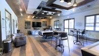 This single-floor amenity space showcases a compelling industrial-modern aesthetic, blending raw materials with contemporary design elements. Exposed brick walls serve as a defining architectural feature, complemented by corrugated metal ceiling panels and a bold grid of dark steel beams overhead. Pendant-style industrial lights hang at varying heights, providing warm, focused illumination throughout the room.

The space is organized into distinct activity zones. A central billiards table anchors the room, surrounded by high-top bar tables and sleek black chairs, creating an ideal setting for casual gatherings. A large flat-screen television is mounted on the brick wall alongside a lounge seating area, while an arcade game station adds an additional entertainment option nearby.

Wide-plank hardwood flooring in a mixed-tone finish runs throughout, adding warmth and grounding the otherwise raw aesthetic. Natural light enters through large windows and a glass door on one side, offering views of an outdoor area with additional seating. The overall ambiance is energetic yet comfortable, making this space well-suited for social interaction and recreational activities.