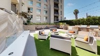 This thoughtfully designed outdoor amenity courtyard offers a versatile and inviting environment ideal for relaxation and entertaining. The space is anchored by a generous turf lawn area furnished with white wicker lounge seating, accent chairs, and coffee tables accented with vibrant orange throw pillows, creating a comfortable and lively atmosphere under open skies. String lights are strung overhead throughout the courtyard, adding a warm, festive ambiance for evening gatherings.

A fully equipped outdoor kitchen station sits along one perimeter, featuring dual stainless steel grills set into a wood-paneled base with a clean white countertop, accompanied by a long communal dining table — perfect for large group cookouts. A built-in bench seating area with colorful cushions lines the raised planter walls, offering additional gathering space surrounded by lush greenery and tropical plantings that provide natural privacy.

A striking chevron-patterned stone accent wall serves as a bold architectural focal point, its veined grey marble-toned tiles adding texture and sophistication to the space. A sleek white ledge runs alongside it, embedded with flush-mounted fixtures. The entire courtyard is framed by the residential building rising above, with palm trees and manicured hedgerows completing the Southern California-inspired aesthetic.