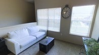 This single-floor space offers a welcoming and well-lit interior designed for everyday comfort. The living area is a standout feature, characterized by large windows fitted with horizontal blinds that allow natural light to filter in while maintaining privacy. The room is finished with soft, wall-to-wall carpeting that adds warmth underfoot and contributes to the overall cozy atmosphere. Neutral-toned walls complement the light and airy feel of the space, creating a versatile backdrop suitable for a variety of décor styles. A white sofa serves as the centerpiece of the living area, paired with a dark vintage-style trunk coffee table that adds a touch of character and contrast. A large decorative wall clock with a classic Roman numeral face adds a distinctive focal point to the room, reinforcing the space's blend of modern simplicity and timeless charm. The layout is open and functional, making efficient use of the available floor space. Overall, this single-story space presents a clean, move-in-ready environment with thoughtful design details that balance practicality with aesthetic appeal.