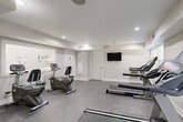 Located at 3901 Campbellton Rd SW in Atlanta, Georgia, this fitness center offers a well-appointed exercise space designed for efficient, functional workouts. The room features a neutral color palette of soft grays and crisp whites, creating a clean, modern atmosphere conducive to focused exercise.

The space is equipped with multiple treadmills and recumbent exercise bikes, providing options for both cardiovascular and low-impact training. A wall-mounted flat-screen television is positioned for easy viewing during workouts. A frosted-glass interior window bearing the property's branding adds a polished, professional touch to the space.

Recessed ceiling lighting ensures the room is well-lit throughout, complemented by natural light entering through windows along one wall. The durable, textured gray flooring is suited for fitness use and easy maintenance. The room includes multiple entry and exit points, with a glass-panel exterior door providing access to the outdoors and an additional interior door for convenient connectivity within the building.

HVAC vents are integrated into the ceiling and walls, maintaining a comfortable environment year-round. The overall layout is thoughtfully organized, allowing adequate space between equipment for safe and comfortable use. This fitness center serves as a practical, well-maintained amenity for residents of the community.