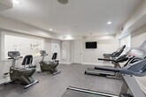 Located at 3901 Campbellton Rd SW in Atlanta, Georgia, this fitness center offers a well-appointed exercise space designed for efficient, functional workouts. The room features a neutral color palette of soft grays and crisp whites, creating a clean, modern atmosphere conducive to focused exercise.

The space is equipped with multiple treadmills and recumbent exercise bikes, providing options for both cardiovascular and low-impact training. A wall-mounted flat-screen television is positioned for easy viewing during workouts. A frosted-glass interior window bearing the property's branding adds a polished, professional touch to the space.

Recessed ceiling lighting ensures the room is well-lit throughout, complemented by natural light entering through windows along one wall. The durable, textured gray flooring is suited for fitness use and easy maintenance. The room includes multiple entry and exit points, with a glass-panel exterior door providing access to the outdoors and an additional interior door for convenient connectivity within the building.

HVAC vents are integrated into the ceiling and walls, maintaining a comfortable environment year-round. The overall layout is thoughtfully organized, allowing adequate space between equipment for safe and comfortable use. This fitness center serves as a practical, well-maintained amenity for residents of the community.