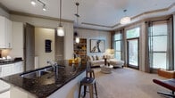 This single-floor residence showcases a thoughtfully designed open-concept layout that seamlessly connects the kitchen, dining, and living areas. The kitchen is a standout feature, equipped with stainless steel appliances, white shaker cabinetry, a dark granite peninsula with an undermount sink, a tile backsplash, and pendant lighting overhead. The peninsula doubles as a breakfast bar with seating, making it ideal for casual dining or entertaining.

The living room is generously proportioned, highlighted by a tray ceiling with crown molding and an elegant chandelier. Large windows allow ample natural light to fill the carpeted space, offering views of the outdoors. The room also features a media area with a wall-mounted television and an arched architectural detail that adds character.

The bathroom is well-appointed with a double-sink granite vanity, white cabinetry, tile flooring, and a tub-shower combination. A dedicated laundry closet tucked between the kitchen and living areas provides added convenience, housing a full-size washer and dryer behind paneled doors. Throughout the residence, consistent use of neutral tones, tile flooring in wet areas, and quality finishes contribute to a cohesive and polished aesthetic.