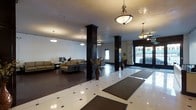 This single-floor lobby presents a polished and welcoming environment defined by a harmonious blend of classic and contemporary design elements. The expansive floor plan is anchored by striking dark-finished structural columns that draw the eye upward toward a smooth, light-colored ceiling adorned with a series of elegant pendant light fixtures featuring warm-toned glass shades and brass hardware, casting a soft, inviting glow throughout the space.

The flooring consists of large-format white tiles accented with dark inlay details, lending a refined, geometric pattern underfoot. Entry mats are positioned near the main entrance, which features full-height glass doors that allow natural light to flow into the interior and offer views to the exterior.

A comfortable seating area is arranged to one side, furnished with neutral-toned sofas and low-profile chairs, providing a relaxed gathering space for visitors. Decorative floor vases with dried botanical arrangements are placed throughout, adding organic warmth to the otherwise structured aesthetic.

The overall ambiance is that of a well-maintained, professionally designed communal space — open, bright, and functional — with thoughtful touches that balance utility with visual appeal. The layout promotes easy circulation, making it an accessible and practical entry point for all who pass through.