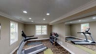 This dedicated fitness space is thoughtfully designed to support a comprehensive workout experience. The room features a textured ceiling adorned with recessed lighting, which provides bright, even illumination throughout the space. Crown molding and neutral warm-toned walls lend the room a clean, polished appearance that distinguishes it from a typical gym environment.

The flooring is covered in a durable patterned carpet with earthy tones, offering both comfort and traction during exercise. A treadmill is positioned toward the front of the room, providing a dedicated space for cardio training. Along one wall, a well-organized rack holds an extensive collection of dumbbells and kettlebells in varying weights, making strength training highly accessible. Exercise mats are neatly laid out on the floor, designating an area for stretching, yoga, or floor-based workouts.

A large wall mirror is mounted on one side of the room, serving both a functional and spatial purpose — allowing users to monitor their form while visually expanding the room. A window allows natural light to filter in, adding a refreshing brightness to the space. The overall layout is practical and efficient, making productive use of the available area while maintaining an organized and welcoming atmosphere for fitness activities.