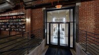 Located at 220 S 47th St in West Philadelphia, this striking industrial space offers a commanding single-floor layout with a substantial open-plan footprint. The architectural design draws heavily from its industrial roots, showcasing exposed brick walls, raw concrete mushroom-cap columns, and a distinctive coffered concrete ceiling with visible ductwork and conduit running throughout — all contributing to an authentic warehouse aesthetic.

Polished concrete floors reflect the carefully curated pendant and track lighting above, creating a warm yet industrial ambiance across the entire space. The main area is divided into distinct functional zones: a central lounge features deep leather Chesterfield sofas and modern accent chairs arranged around a coffee table; a conference and dining area offers a long communal table with metal chairs suited for meetings or events; and a recreation zone includes a foosball table, ping pong table, and pool table.

Additional areas include a library nook with floor-to-ceiling bookshelves, private room enclosures along the perimeter, and a reception or service counter. The entry features a glass double-door vestibule with exposed brick, steel railings, and an accessible ramp. Throughout, the space balances raw, industrial character with purposeful furnishings, making it a versatile venue for events, collaboration, and community gatherings.