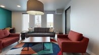 Located at 3250 Hollis St in Oakland, California, this single-floor community space offers a thoughtfully designed environment that blends comfort with functionality. The layout is divided into two distinct yet connected zones, seamlessly linked by glass-panel interior doors with sleek black steel frames.

The main lounge area is anchored by large floor-to-ceiling windows that flood the room with natural light and frame views of the surrounding Oakland neighborhood. A bold, colorful geometric area rug defines the seating arrangement, which features a tufted charcoal sofa, a rust-red loveseat, and a low-profile wooden coffee table. A wall-mounted split air conditioning unit ensures year-round comfort, and a statement pendant lamp adds warmth overhead. Light-toned herringbone hardwood flooring runs throughout, adding a sophisticated, modern touch.

The adjoining co-working area is energized by a striking teal accent wall adorned with a curated gallery of framed abstract artwork. Two dedicated wooden desks with rolling chairs provide focused workstations, while the rust-red sofa creates a relaxed seating option nearby. A decorative patterned console completes the space. Recessed ceiling lighting ensures even illumination throughout. Together, these two areas create a vibrant, well-appointed communal space ideal for both collaboration and relaxation.