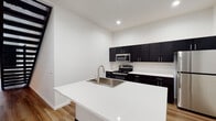 This two-floor space showcases a clean, modern design defined by a crisp white-and-dark contrast palette throughout. The kitchen serves as a central highlight, featuring dark espresso cabinetry extending to the upper walls, white quartz countertops, stainless steel appliances including an electric range and over-the-range microwave, and a freestanding island with an integrated sink. Light-toned hardwood-style flooring runs through the main living areas, adding warmth to the contemporary aesthetic.

The upper level is distinguished by striking vaulted ceilings with exposed dark beams, creating an open, loft-like atmosphere. Carpeted bedroom spaces on both floors provide comfort, with one bedroom featuring a window offering views of surrounding greenery. A dedicated in-unit laundry area with a full washer and dryer adds everyday convenience.

The bathroom is well-appointed with dark cabinetry, a dual-sink vanity with a large mirror, wood-look flooring, and a walk-in shower. A second bathroom includes a single-sink vanity with matching cabinetry. Recessed lighting and clean white walls throughout contribute to the space's bright, minimalist ambiance.