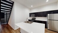 This two-floor space showcases a clean, modern design defined by a crisp white-and-dark contrast palette throughout. The kitchen serves as a central highlight, featuring dark espresso cabinetry extending to the upper walls, white quartz countertops, stainless steel appliances including an electric range and over-the-range microwave, and a freestanding island with an integrated sink. Light-toned hardwood-style flooring runs through the main living areas, adding warmth to the contemporary aesthetic.

The upper level is distinguished by striking vaulted ceilings with exposed dark beams, creating an open, loft-like atmosphere. Carpeted bedroom spaces on both floors provide comfort, with one bedroom featuring a window offering views of surrounding greenery. A dedicated in-unit laundry area with a full washer and dryer adds everyday convenience.

The bathroom is well-appointed with dark cabinetry, a dual-sink vanity with a large mirror, wood-look flooring, and a walk-in shower. A second bathroom includes a single-sink vanity with matching cabinetry. Recessed lighting and clean white walls throughout contribute to the space's bright, minimalist ambiance.