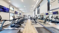 Located at 420 N Dean Rd in Auburn, Alabama, this well-equipped fitness center offers a single-floor layout designed to support a comprehensive workout experience. The space is defined by a long, open floor plan that maximizes usability, with a clean and energetic aesthetic throughout.

The flooring combines light hardwood and patterned geometric carpet tiles, creating distinct zones for cardio and strength training. A row of True-brand treadmills, ellipticals, and stationary bikes lines one side of the room, while cable machines, a multi-function strength station, free weights, and a weight bench occupy the opposing side. Full-length mirrors along one wall expand the sense of space and support proper form during workouts.

Bold orange accent walls punctuate the room and feature motivational phrases, contributing to an upbeat and energizing atmosphere. Multiple flat-screen TVs are mounted throughout, allowing users to stay entertained during workouts. Recessed ceiling lighting combined with numerous windows along the walls allows ample natural light to fill the space, with views toward an outdoor pool area. A water fountain is also available within the room for convenience.

The overall design is modern, functional, and inviting — a well-appointed facility suited for a wide range of fitness routines.