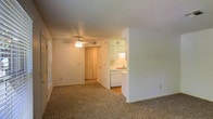 This single-floor space offers a practical and functional layout designed for comfortable everyday living. The main living area features wall-to-wall carpeting that adds warmth underfoot, while a ceiling fan with a light fixture provides both air circulation and overhead lighting. A textured ceiling runs throughout, lending a classic residential character to the space. The living area flows naturally toward a set of interior doors, suggesting access to additional rooms beyond. The kitchen is positioned adjacent to the living space and is accessible via an open pass-through, promoting a connected feel between the two areas. It is equipped with white cabinetry providing ample storage, white appliances including a dishwasher, and a sink. The kitchen floor transitions to a hard surface, contrasting with the carpeted living area. Neutral white walls throughout the space create a clean, bright backdrop that allows for flexible interior styling. Horizontal blinds on the windows offer light control and privacy. Overall, the layout is straightforward and efficient, making it well-suited for those seeking a low-maintenance, move-in-ready living environment.