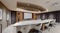 Located at 99 Wilkens Ln in Barnstable, MA, this polished single-floor presentation and screening room offers a refined, theater-inspired environment designed for professional gatherings and screenings. The space is defined by its striking curved, tiered layout, which organizes seating across two distinct levels for optimal sightlines to the large projection screen at the front.

The front tier features a sweeping marble-topped work surface with modern patterned swivel chairs, providing a functional and stylish workspace. Behind it, the upper tier is furnished with rich cognac leather club chairs arranged in curved rows, offering a comfortable viewing area. The focal wall showcases warm wood paneling flanking the projection screen, accented by decorative wall sconces and textured tile details that add depth and visual interest.

The coffered ceiling design features a dramatic curved tray detail with recessed lighting, lending an intimate yet elevated ambiance to the room. Full-length charcoal drapery lines the walls, enhancing acoustics while contributing to the space's sleek, contemporary aesthetic. Patterned carpet, curated artwork on the walls, and a secondary service counter with decorative vessels complete the sophisticated interior. The overall design balances professional functionality with upscale, hospitality-inspired style.