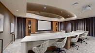 Located at 99 Wilkens Ln in Barnstable, MA, this polished single-floor presentation and screening room offers a refined, theater-inspired environment designed for professional gatherings and screenings. The space is defined by its striking curved, tiered layout, which organizes seating across two distinct levels for optimal sightlines to the large projection screen at the front.

The front tier features a sweeping marble-topped work surface with modern patterned swivel chairs, providing a functional and stylish workspace. Behind it, the upper tier is furnished with rich cognac leather club chairs arranged in curved rows, offering a comfortable viewing area. The focal wall showcases warm wood paneling flanking the projection screen, accented by decorative wall sconces and textured tile details that add depth and visual interest.

The coffered ceiling design features a dramatic curved tray detail with recessed lighting, lending an intimate yet elevated ambiance to the room. Full-length charcoal drapery lines the walls, enhancing acoustics while contributing to the space's sleek, contemporary aesthetic. Patterned carpet, curated artwork on the walls, and a secondary service counter with decorative vessels complete the sophisticated interior. The overall design balances professional functionality with upscale, hospitality-inspired style.