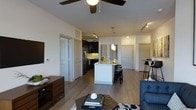This thoughtfully designed single-floor residence showcases an open-concept layout that effortlessly connects the main living areas. The living room offers a welcoming atmosphere anchored by a ceiling fan with light fixture and wood-look flooring that flows throughout the space, adding warmth and visual continuity.

The kitchen stands out as a focal point of the home, featuring dark cabinetry, light-colored countertops, and stainless steel appliances including a refrigerator. A central white kitchen island with bar seating provides both additional workspace and casual dining options, illuminated by elegant pendant lights hanging above. Track lighting accents the kitchen area, ensuring bright and functional illumination.

Adjacent to the kitchen, a dining area with bar-height seating offers an additional space for meals or entertaining. The open sightlines between the kitchen, dining, and living zones create a sense of spaciousness and make the layout ideal for both daily living and hosting guests.

The space features multiple doors leading to additional rooms, with white interior doors and trim providing a clean, modern aesthetic against the neutral-toned walls. The overall ambiance is bright and contemporary, combining functional design with stylish finishes to create a comfortable and inviting home environment.