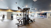 Located at 1220 South Ave in Plainfield, NJ, this generously sized fitness center offers a comprehensive and thoughtfully designed exercise environment across a single floor. The space is organized to accommodate a wide variety of workout styles, from cardiovascular training to strength and functional fitness.

The main exercise area is expansive, featuring durable rubberized flooring throughout and dark, stylish ceilings fitted with recessed lighting and multiple ceiling fans to maintain airflow and comfort. Large floor-to-ceiling windows and sliding glass doors line one side of the space, flooding the interior with natural light and offering views of the surrounding outdoor area.

Equipment is plentiful and well-arranged, including a row of treadmills, cable and pulley machines, a power rack with weight plates, kettlebells, medicine balls, stability balls, and a dedicated stretching and mat area. A built-in shelving and storage unit provides organized space for accessories and personal items.

A striking geometric mural in bold, multicolored tones serves as a vibrant focal point on one wall, complemented by a large mounted flat-screen television. Arched doorways and clean architectural lines add character to the overall design. The fitness center also benefits from a dedicated free weight and functional training zone, making it a fully equipped destination for all fitness levels.