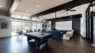 This contemporary amenity lounge is designed to deliver a full-service social and entertainment experience across a single, open-plan floor. The space is anchored by a dedicated home theater area, where a large projection screen mounted against a bold, dark accent wall is paired with plush seating — including a deep blue sofa and white armchairs — arranged on a vibrant chevron area rug over rich dark hardwood flooring. Ceiling fans with industrial flair and recessed lighting contribute to the comfortable ambiance throughout.

Adjacent to the theater zone, a full-size billiards table commands the central floor space, providing a natural focal point for social activity. The open layout allows these entertainment zones to flow seamlessly into one another.

A compact but fully appointed kitchenette offers stainless steel appliances — including a full-size refrigerator, wall oven, and microwave — set against striking dark brick tile backsplash and light cabinetry with modern hardware, ideal for hosting events.

The lounge also features a bright café-style seating area lined with floor-to-ceiling windows that flood the space with natural light and frame views of the surrounding grounds and pool area. The grand entry corridor, with statement pendant lighting and decorative wall paneling, sets an impressive tone upon arrival.