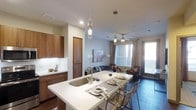 This single-floor residence is designed around an open-concept layout that seamlessly connects the kitchen, dining, and living areas into one cohesive, functional space. The kitchen features warm wood cabinetry with sleek hardware, a white quartz waterfall-edge island that doubles as a breakfast bar with seating for multiple guests, and an undermount sink with a modern gooseneck faucet. Pendant lighting with cylindrical glass fixtures hangs above the island, adding a refined, contemporary touch.

The dining and living areas share a unified space illuminated by a ceiling fan with integrated lighting and large windows dressed with neutral curtains, allowing generous natural light to fill the room. A secondary door provides additional access, further enhancing the airy feel. Dark hardwood-style flooring runs throughout, providing a rich contrast to the light-colored walls and white ceiling. The living area accommodates comfortable seating arrangements, completing the open, modern aesthetic. The overall ambiance is clean, well-lit, and thoughtfully designed for everyday functionality and entertaining.