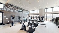 Located at 2510 W 27th Ave in Denver's vibrant LoHi neighborhood, this contemporary fitness center offers a spacious, single-floor layout designed to accommodate a full range of workout needs. The space is defined by soaring double-height ceilings and floor-to-ceiling windows along multiple walls, allowing generous natural light to flood the interior while providing open views of the surrounding Denver cityscape.

The cardio zone features a row of treadmills with individual screens, alongside stationary bikes, StairMaster climbers, rowing machines, and elliptical trainers — all positioned to take advantage of the exterior views. A dedicated strength training area includes a cable and functional training rig, a full dumbbell rack, adjustable benches, and a wall-length mirror that visually expands the space.

The design aesthetic is modern and refined, with light-toned flooring, linear recessed LED lighting accents in the ceiling, and a bold, large-scale abstract wallcovering that adds visual interest throughout. Additional accessories such as medicine balls and resistance equipment are neatly organized on shelving units. Glass doors provide access to an adjacent outdoor area, further enhancing the open, airy character of the facility. The overall ambiance is clean, energetic, and well-appointed.