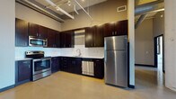 This urban loft blends industrial character with contemporary finishes across a single, well-appointed floor. The kitchen is a standout feature, showcasing rich dark cabinetry, a full suite of stainless steel appliances including a refrigerator, range, built-in microwave, and dishwasher, complemented by a white subway tile backsplash and light countertops. Exposed ductwork and track lighting suspended from the ceiling reinforce the industrial aesthetic throughout.

The living and sleeping areas feature polished concrete floors that reflect natural light, while white-painted brick walls add texture and a sense of history to the space. Large windows with dark frames invite generous amounts of natural light and offer views of the surrounding urban streetscape, enhancing the open, airy atmosphere of each room.

The space includes two bathrooms: a full bath equipped with a tub-shower combination, and a half bath featuring dark cabinetry with a vanity mirror and modern fixtures. Neutral gray tones throughout the bathrooms maintain a clean, contemporary look. The overall design balances raw architectural elements with modern amenities, creating a distinctive and functional living environment with strong urban loft character.