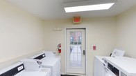 This single-story laundry facility offers a clean, functional space designed to meet residents' everyday laundering needs. The room features light-colored walls and bright overhead fluorescent lighting, including a skylight-style panel that enhances visibility throughout the space. Multiple top-loading washing machines are positioned along the walls, with additional units visible on the opposite side of the room, providing ample capacity for shared use. Safety is a clear priority, as evidenced by the prominently mounted fire extinguisher, a clearly marked exit sign above the door, and a fire alarm panel on the adjacent wall. A glass-paneled exit door allows natural light to filter into the space while offering a view of the surrounding outdoor walkway and neighboring brick building. The door is equipped with a secure electronic lock, ensuring controlled access. The overall layout is straightforward and practical, prioritizing ease of use and circulation within the space. The facility maintains a tidy, well-lit environment suited for communal use.