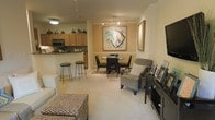 This single-floor residence offers an inviting open-concept layout that effortlessly blends the living, dining, and kitchen areas into one cohesive and functional space. The living area is anchored by plush carpeting in a soft neutral tone, complemented by comfortable seating and a woven ottoman, creating a relaxed yet refined atmosphere. A striking large-scale abstract artwork adorns the wall, serving as a bold focal point that adds character and visual interest to the room. The dining area features a round glass-top table surrounded by dark wood chairs, perfectly positioned beneath a framed art piece that echoes the space's curated aesthetic. Adjacent to the dining and living areas, the kitchen is defined by warm honey-toned cabinetry, a decorative mosaic tile backsplash, and modern stainless steel appliances. A raised breakfast bar with stylish bar stools provides additional seating and naturally delineates the kitchen from the living space. Track lighting runs along the ceiling, ensuring the entire open area is well-illuminated. The overall ambiance is warm, sophisticated, and welcoming, with a cohesive color palette of creams, tans, and dark wood accents tying every zone of the space together seamlessly.