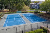 Located in Alpharetta, Georgia, Belmont at Park Bridge offers a well-appointed set of outdoor community amenities designed for active recreation and relaxed outdoor living. The centerpiece of the recreational area is a full-size tennis court with a blue surface and regulation net, enclosed by black iron fencing and illuminated by court lighting for evening play. The court is framed by a lush backdrop of tall mature trees, providing a natural and scenic setting. Adjacent to the tennis court, a swimming pool is visible beyond the iron perimeter fencing, outfitted with poolside lounge seating and vibrant red umbrellas for shade. Landscaped planting beds featuring neatly trimmed hedges, crape myrtle trees, and seasonal flowering plants line the walkways throughout the grounds. Raised retaining walls constructed of timber define tiered landscape areas, adding structure and visual interest to the outdoor environment. Concrete pathways with black iron railings connect the various amenity areas, ensuring accessibility throughout. The community is currently undergoing improvements, as indicated by signage on the property. Overall, the grounds reflect a well-maintained, green, and community-focused outdoor environment suited for residents seeking both active and leisurely outdoor amenities.
