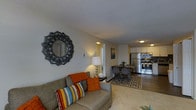 This single-floor residence showcases an open-concept layout that seamlessly connects the living, dining, and kitchen areas into one cohesive space. The living area is anchored by a comfortable grey sofa dressed with colorful throw pillows and an orange accent cushion, complemented by a decorative sunburst mirror mounted on the neutral-toned wall. A small side table with a table lamp adds warmth to the corner.

The space flows naturally into a compact dining area, where a dark-toned table and chairs rest atop a patterned area rug that defines the zone. Beyond this, the kitchen is efficiently laid out with white cabinetry, under-cabinet lighting, and stainless steel appliances including a refrigerator and range. A tile backsplash adds a decorative element to the cooking space.

The flooring transitions from carpeted living areas to hard-surface flooring near the dining and kitchen zones. A textured ceiling runs throughout, and warm overhead lighting illuminates each area. A hallway visible from the living room suggests additional private spaces within the unit. Overall, the layout is practical and well-appointed.