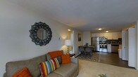 This single-floor residence showcases an open-concept layout that seamlessly connects the living, dining, and kitchen areas into one cohesive space. The living area is anchored by a comfortable grey sofa dressed with colorful throw pillows and an orange accent cushion, complemented by a decorative sunburst mirror mounted on the neutral-toned wall. A small side table with a table lamp adds warmth to the corner.

The space flows naturally into a compact dining area, where a dark-toned table and chairs rest atop a patterned area rug that defines the zone. Beyond this, the kitchen is efficiently laid out with white cabinetry, under-cabinet lighting, and stainless steel appliances including a refrigerator and range. A tile backsplash adds a decorative element to the cooking space.

The flooring transitions from carpeted living areas to hard-surface flooring near the dining and kitchen zones. A textured ceiling runs throughout, and warm overhead lighting illuminates each area. A hallway visible from the living room suggests additional private spaces within the unit. Overall, the layout is practical and well-appointed.