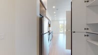 This single-floor unit showcases a clean, contemporary design defined by an open-concept layout and a cohesive neutral palette throughout. Light hardwood flooring extends across the main living areas, complementing the crisp white walls and creating a bright, airy atmosphere. The living area is generously proportioned, featuring floor-to-ceiling windows and a glass sliding door that flood the space with natural light while offering views of surrounding greenery. A wall-mounted mini-split system provides efficient climate control.

The kitchen is well-appointed with warm wood-toned cabinetry extending to the ceiling, a full suite of stainless steel appliances including a gas range, microwave, dishwasher, and refrigerator, as well as a single-basin sink with modern fixtures. A closet adjacent to the kitchen provides additional storage.

The bathroom maintains the modern aesthetic with large-format light tile flooring, a wood-toned vanity topped with a white countertop, a backlit mirror, and a glass-enclosed tub-shower combination. Recessed lighting throughout the unit enhances the clean, uncluttered feel of the overall space.