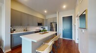 This thoughtfully designed single-floor residence showcases a cohesive contemporary aesthetic throughout, with rich hardwood flooring that flows seamlessly across every room. The kitchen is a standout feature, outfitted with grey shaker-style cabinetry, crisp white quartz countertops, a classic subway tile backsplash, and a full suite of stainless steel appliances including a gas range, dishwasher, microwave, and refrigerator. A generous island with seating provides both additional prep space and a casual dining area, making the kitchen as functional as it is visually appealing. The striking teal entry door adds a bold, welcoming touch to the space. The open living area is bathed in natural light, courtesy of expansive floor-to-ceiling windows that frame outdoor views and lend an airy, connected feel to the interior. The bedroom continues the modern design language, featuring a large black-framed window that fills the room with natural light and offers pleasant outdoor views. The bathroom is finished with grey cabinetry, a clean white vanity, and warm hardwood-style flooring, maintaining the consistent, polished palette found throughout the residence. Overall, the space presents a clean, modern design with carefully selected finishes and a bright, welcoming atmosphere.
