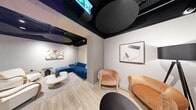 This thoughtfully designed single-floor space offers a compelling blend of industrial-chic architecture and contemporary interior design. The exposed black ceiling reveals ductwork, structural elements, and dramatic circular acoustic panels, creating a bold visual contrast against the light wood flooring and neutral-toned walls that run throughout.

The space is organized into several distinct zones. A central lounge area features intimate seating arrangements, including a vibrant blue sectional sofa, warm terracotta and cream armchairs, and a sculptural white coffee table, all anchored by curated abstract artwork on the walls. Light wood partition columns subtly divide the open floor plan into connected but defined pockets, enhancing privacy while maintaining flow.

A second lounge zone offers elegant white and gold armchairs alongside a sleek black side table, reflecting a more refined aesthetic. A dedicated meeting or dining room accommodates a large dark marble-topped table surrounded by upholstered seating — ideal for collaborative gatherings or formal discussions.

A galley-style kitchen rounds out the space, featuring navy blue cabinetry, white countertops, a textured tile backsplash, bar seating, and a striking terrazzo floor. Recessed and track lighting throughout ensures each area is well-illuminated. The overall ambiance is polished, modern, and versatile.