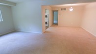 This single-floor residence features an open, carpeted layout with a clean, neutral aesthetic. The main living area is finished with wall-to-wall beige carpet and light-toned walls, creating a bright and airy atmosphere. A pendant light fixture hangs from the ceiling, providing focused overhead illumination to the space. A dedicated laundry nook is conveniently integrated into the floor plan, housing a stacked washer and dryer unit tucked behind a partial wall — a practical feature that adds significant everyday convenience. A visible HVAC vent is positioned near the ceiling, ensuring climate control throughout. The open sightline from the main area leads to an additional room beyond, suggesting a functional layout with well-connected spaces. White baseboards trim the perimeter of the rooms, adding a subtle architectural finish. The overall design is straightforward and functional, with an emphasis on open space and ease of movement between areas.