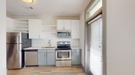 This single-floor unit offers a clean, contemporary design with a neutral color palette throughout. The kitchen is a standout feature, outfitted with two-tone cabinetry — white upper cabinets with open shelving and gray lower cabinets — complemented by a white subway tile backsplash, light countertops, and a full suite of stainless steel appliances including an electric range, over-the-range microwave, and dishwasher. A single-basin sink and large window with blinds allow natural light to fill the space. Wood-look flooring runs throughout the main living and kitchen areas, extending into the hallway, which leads from the kitchen toward a windowed entry door, creating a bright, airy corridor. One room features soft carpeting, a ceiling fan, and a wall-mounted electrical panel, providing a flexible living or sleeping area. An additional room showcases the same wood-look flooring with a large window that floods the space with natural light, ideal as a bedroom or office. The bathroom is accessed via the hallway and features wood-look tile flooring, a dark-cabinet vanity with a granite-style countertop, and clean white walls. The overall design is modern, functional, and well-lit throughout.