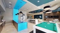 This expansive community lounge is a thoughtfully designed social hub that seamlessly blends entertainment, relaxation, and productivity. The interior is defined by a striking contemporary aesthetic, anchored by bold teal ceiling panels and illuminated blue-backlit glass partitions that create vivid focal points throughout the space. Large-format porcelain tile flooring runs throughout, lending a polished, cohesive look.

At the heart of the lounge, a sleek white pool table invites recreation, while a bar area features illuminated blue countertops, clear acrylic stools, pendant lighting, and an integrated TV — ideal for casual gatherings. A prep kitchen with stainless steel cabinetry and white countertops supports entertaining needs.

The lounge area offers comfortable seating arrangements positioned near a dramatic dark decorative wall feature, complemented by multiple flat-screen TVs and warm wood-paneled accent walls. A dedicated workspace includes computer stations and organized shelving alongside a glass-enclosed library nook.

Floor-to-ceiling windows dressed with patterned sheer curtains flood the space with natural light and offer views of the surrounding grounds, while bistro-height tables provide intimate seating near the glass doors leading to the outdoor terrace.