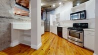 Located at 3502 Scotts Ln in Philadelphia's 19129 zip code, Unit 822 is a single-floor loft-style residence that masterfully combines historic industrial character with contemporary updates. The generously sized floor plan provides ample living space across an open, flowing layout.

The living area is a standout, showcasing polished wide-plank pine hardwood floors, exposed heavy timber ceiling beams, and original stone masonry walls that speak to the building's historic origins. Structural concrete columns punctuate the space, adding to its authentic industrial aesthetic. Exposed ductwork runs overhead, complementing the raw architectural details throughout.

The kitchen features white cabinetry with open shelving, stainless steel appliances including a gas range and refrigerator, and a dark sink fixture. The concrete ceiling and pine floors carry through seamlessly, maintaining visual continuity.

Additional rooms include well-proportioned spaces with exposed beam ceilings and horizontal windows that bring in natural light. Two closets provide practical storage. The two bathrooms contrast the industrial common areas with clean, refined finishes — one features large-format beige floor tiles, a soaking tub with tile surround, and a frameless glass-enclosed walk-in shower with a built-in bench and recessed shelving.