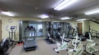 This fitness center is designed to support a well-rounded cardio workout routine in a comfortable and functional setting. The space is outfitted with multiple pieces of exercise equipment, including treadmills, elliptical trainers, and stationary bikes, thoughtfully arranged to allow for ease of movement between machines. Large floor-to-ceiling glass sliding doors line one wall, flooding the room with natural light and providing views to the outdoors, creating an open and inviting atmosphere. Full-length mirrors along the adjacent wall further enhance the sense of space while allowing users to monitor their form during workouts. The textured ceiling features recessed fluorescent lighting alongside a ceiling fan, ensuring the room remains well-lit and ventilated during use. Rubber floor mats beneath the equipment offer stability and protect the flooring. A wall clock keeps exercisers on track, and the overall layout is clean and organized. The room's neutral color palette and practical design make it a focused, distraction-free environment well-suited for daily fitness activity.