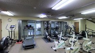 This fitness center is designed to support a well-rounded cardio workout routine in a comfortable and functional setting. The space is outfitted with multiple pieces of exercise equipment, including treadmills, elliptical trainers, and stationary bikes, thoughtfully arranged to allow for ease of movement between machines. Large floor-to-ceiling glass sliding doors line one wall, flooding the room with natural light and providing views to the outdoors, creating an open and inviting atmosphere. Full-length mirrors along the adjacent wall further enhance the sense of space while allowing users to monitor their form during workouts. The textured ceiling features recessed fluorescent lighting alongside a ceiling fan, ensuring the room remains well-lit and ventilated during use. Rubber floor mats beneath the equipment offer stability and protect the flooring. A wall clock keeps exercisers on track, and the overall layout is clean and organized. The room's neutral color palette and practical design make it a focused, distraction-free environment well-suited for daily fitness activity.