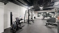 Located at 856 Gramercy Dr in Los Angeles, this dedicated exercise room delivers a fully outfitted fitness experience within a compact yet efficiently organized single-floor layout. The space features a striking industrial-modern aesthetic, with exposed ceiling ductwork and piping set against a dark ceiling, white concrete block walls, and durable rubber flooring throughout — a combination that is both functional and visually cohesive.

The strength training area is well-appointed, housing a multi-functional cable machine, a Smith machine, a leg press, adjustable benches, and a variety of free weight and resistance equipment. The cardio section includes treadmills and a stair climber, providing options for varied workout routines. Large wall-mounted mirrors span multiple sections of the room, enhancing the sense of space and allowing users to monitor their form during exercise.

Recessed and overhead lighting ensures the entire floor is evenly illuminated, while a glass-panel partition wall with black metal framing adds a modern architectural touch and visually separates zones within the space. Structural columns are integrated cleanly into the layout without disrupting the flow of the floor. The overall ambiance is focused and professional, making this exercise room a capable and well-designed fitness destination.