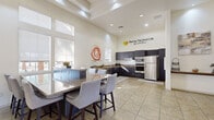 Located at 7606 W Peoria Ave in Peoria, Arizona, this single-floor community space is home to the Sharing The Good Life Foundation. The open-concept layout seamlessly integrates a fully equipped kitchen and a generous dining area, creating a welcoming and functional environment suited for group gatherings and community programming.

The kitchen is outfitted with sleek dark cabinetry, stainless steel appliances including a full-size refrigerator, double wall oven, microwave, and dishwasher, along with ample counter space and a dedicated sink area. Vibrant wall art adds a pop of color and warmth to the culinary space.

The dining area centers around a large granite-topped communal table surrounded by upholstered counter-height chairs, comfortably seating a sizeable group. Oversized windows dressed with roller shades flood the space with natural light, while soaring ceilings with a coffered detail enhance the sense of openness and volume.

Light-toned tile flooring runs throughout, contributing to the bright, airy atmosphere. A connected entry corridor features additional seating, a wall-mounted television, and glass doors that invite natural views of the surrounding Peoria landscape. The overall ambiance is clean, modern, and purposefully designed to foster community engagement.