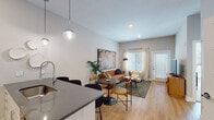 Located along Berkley Pkwy in Kansas City, MO, this single-floor unit offers a thoughtfully designed open-concept layout that seamlessly connects the kitchen, dining, and living areas. The kitchen is well-appointed with white flat-front cabinetry extending to the ceiling, stainless steel appliances including a French-door refrigerator, electric range, and built-in microwave, all complemented by dark quartz countertops and a undermount sink. Globe pendant lights hang above the kitchen peninsula, which doubles as a casual dining counter with bar seating.

The open living and dining area features light hardwood flooring, neutral gray walls, and large windows dressed with sheer curtains and horizontal blinds, allowing generous natural light throughout. A private entrance door with sidelight windows adds convenience and additional brightness to the space.

The bedroom is carpeted and features a large multi-pane window offering outdoor views, with a ceiling fan providing year-round comfort. The full bathroom is clean and functional, finished with tile surround, a soaking tub-shower combination, hardwood-style flooring, and a white vanity. The overall aesthetic throughout the unit is modern and cohesive, with a warm, contemporary ambiance.
