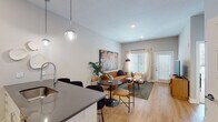 Located along Berkley Pkwy in Kansas City, MO, this single-floor unit offers a thoughtfully designed open-concept layout that seamlessly connects the kitchen, dining, and living areas. The kitchen is well-appointed with white flat-front cabinetry extending to the ceiling, stainless steel appliances including a French-door refrigerator, electric range, and built-in microwave, all complemented by dark quartz countertops and a undermount sink. Globe pendant lights hang above the kitchen peninsula, which doubles as a casual dining counter with bar seating.

The open living and dining area features light hardwood flooring, neutral gray walls, and large windows dressed with sheer curtains and horizontal blinds, allowing generous natural light throughout. A private entrance door with sidelight windows adds convenience and additional brightness to the space.

The bedroom is carpeted and features a large multi-pane window offering outdoor views, with a ceiling fan providing year-round comfort. The full bathroom is clean and functional, finished with tile surround, a soaking tub-shower combination, hardwood-style flooring, and a white vanity. The overall aesthetic throughout the unit is modern and cohesive, with a warm, contemporary ambiance.