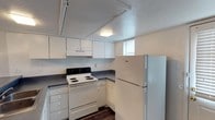 This two-floor space offers a practical and straightforward layout designed for functional living. The kitchen is a notable feature, outfitted with white cabinetry that lines the upper and lower walls, providing ample storage. Dark countertops create a subtle contrast against the white cabinets and appliances. Essential appliances are included, among them a white electric coil range and a full-size refrigerator, making the kitchen move-in ready. A small window positioned near the ceiling allows a modest amount of natural light to filter into the space, supplemented by a ceiling-mounted light fixture overhead. The kitchen floor features dark hardwood-style flooring, adding a touch of warmth to the otherwise neutral palette. A textured ceiling adds subtle visual interest throughout. The two-floor configuration of this space provides a degree of separation between living areas, offering practical versatility. The overall aesthetic is clean and minimal, with a neutral white and grey color scheme that provides a blank canvas adaptable to a range of personal styles and furnishing preferences.