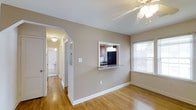 This single-floor residence offers a well-organized layout designed for comfortable everyday living. The main living and dining area features warm hardwood flooring, neutral beige walls, and a ceiling fan that enhances both comfort and airflow. Natural light filters in through a window fitted with blinds, lending the space a bright and airy feel. A distinctive architectural pass-through window with a granite countertop ledge connects the dining area to the kitchen, creating an open and interactive flow between the two spaces.

The kitchen is efficiently designed and well-appointed, showcasing rich wooden cabinetry with modern hardware that provides ample storage. Granite countertops run along the workspace, complemented by a suite of black appliances including a refrigerator and electric range, as well as a stainless steel over-the-range microwave. A window above the countertop brings in additional natural light, making the kitchen feel open despite its compact footprint.

A hallway visible from the main living area leads to additional rooms, including a bathroom with classic white fixtures. White trim and baseboards throughout the home provide clean contrast against the warm-toned walls and flooring, contributing to a cohesive and polished interior aesthetic.