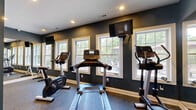 The Covered Bridge Fitness Center at 9 Covered Bridge Rd in Newtown, CT, is a dedicated exercise room designed to support a well-rounded workout routine. The space is finished with deep navy-blue walls and crisp white trim, creating a sleek, focused atmosphere complemented by neutral carpet flooring and recessed lighting throughout.

A row of large, white-framed windows lines one wall, flooding the room with natural light and offering views of an outdoor patio and surrounding greenery. Wall-mounted flat-screen televisions are positioned throughout, providing entertainment during cardio sessions.

The room is outfitted with a selection of Life Fitness cardio equipment, including treadmills, elliptical trainers, and upright stationary bikes. A free-weight rack with dumbbells is also available for strength training. Full-length mirrors along one wall visually expand the space and allow users to monitor their form during exercise.

Sanitation supplies, including a stainless steel wipe dispenser, are conveniently placed near the entrance, reflecting a commitment to cleanliness and user comfort. Signage at the entrance welcomes members and outlines usage guidelines. The Covered Bridge Fitness Center offers a well-appointed, bright, and functional environment suited for a variety of fitness activities.