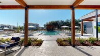 This expansive outdoor amenity space is designed to deliver a resort-like experience within a residential community. At its center sits a generously sized swimming pool with crystal-clear turquoise water, framed by a wide concrete pool deck lined with lounge chairs and shaded by multiple market umbrellas — ideal for relaxing under open skies. A striking pergola-style cabana, constructed with warm-toned wood beams and black metal hardware, anchors one end of the pool area and provides a shaded gathering point with direct sightlines across the water. At the opposite side, a dedicated outdoor kitchen and grilling station offers a functional space for entertaining. The adjacent covered patio, featuring soaring ceilings with exposed wood beams, ceiling fans, and large windows, is furnished with plush navy blue seating and round dining tables, creating a comfortable indoor-outdoor transition zone. Mature oak trees and manicured landscaping with ornamental shrubs and mulched beds add natural character and privacy to the space. The surrounding residential buildings, finished in white with neutral gray accents, complement the clean, modern-farmhouse aesthetic carried throughout the amenity area. The overall ambiance is bright, open, and inviting, with ample space for both social gatherings and quiet relaxation.