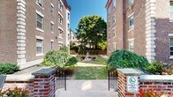 Located at 6803 Emlen Street in Philadelphia's desirable Mt. Airy neighborhood (19119), this multi-story brick building showcases timeless early 20th-century architecture with distinctive limestone quoins, rounded turret corners, and ornate ironwork detailing. The building is set back from the street and anchored by an elegantly designed shared outdoor space that serves as a central gathering area for residents.

The courtyard is a standout feature, framed by the building's classic brick facades on multiple sides. A decorative multi-tiered stone fountain serves as the focal point of the landscaped garden, surrounded by manicured boxwood shrubs, neatly trimmed topiary columns, and mature shade trees. Wrought iron entry gates with decorative scrollwork open onto a spacious stone-paved patio furnished with seating and colorful planters, creating an inviting outdoor retreat.

Additional green space flanks the courtyard, offering grassy lawns with iron benches nestled among perennial plantings and well-maintained garden beds. Stone retaining walls and brick borders define the various landscaped zones, contributing to the cohesive and refined aesthetic. The outdoor areas are bathed in natural light, offering a peaceful urban sanctuary with a strong sense of architectural character and community.